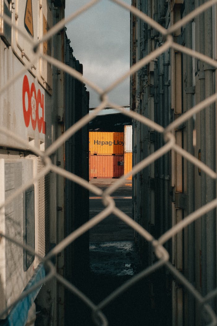 Shipping containers viewed through a chain-link fence in an industrial area.